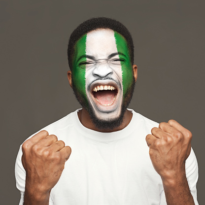 Face of young screaming african-american man painted with flag of Nigeria. Football or soccer team fan, sport event, faceart and patriotism concept. Studio shot at gray background, copy space