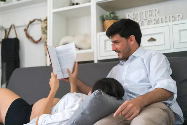 Handsome husband reading book and asian pregnant woman playing tablet computer with lying on sofa in the living room at home. concept of happy family takecare and healthy.