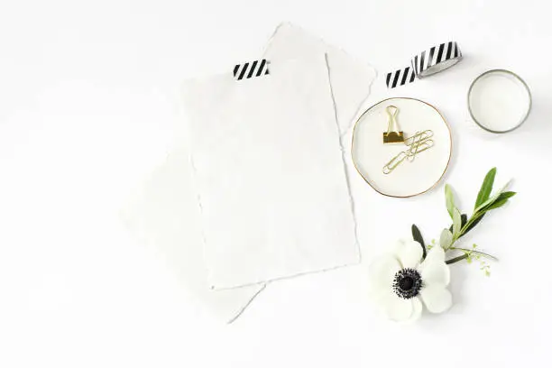 Wedding, birthday desktop mock-up scene. Blank cotton paper greeting cards, washi tape and golden clips. Olive branch and anemone flower, white table background. Flat lay, top view. Feminine stationery.