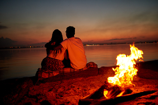 Rear view of romantic couple sitting on log covered with plaid and embracing while contemplating tranquil sea on beach, burning campfire behind them at night