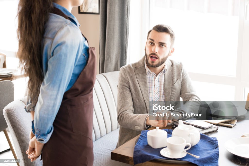 Pensive customer asking waitress in cafe Pensive handsome businessman in light gray jacket sitting at table with teapot and cup and asking waitress about lunch while making order in restaurant Asking Stock Photo