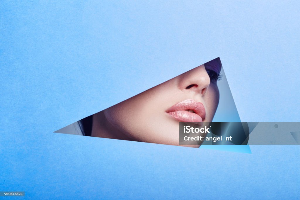 Woman looking in the hole, bright beautiful makeup, big eyes and lips, bright lipstick, professional cosmetics and facial care. Bright colored background and a gap slot in the paper Human Lips Stock Photo