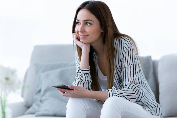 istock Pretty young woman looking sideways while using her mobile phone on sofa at home. 950874694