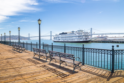 Benches at historic Pier 7 with traditional paddleboat and Oakland Bay Bridge in the background on a sunny day with bliue sky and clouds, San Francisco, California, USA