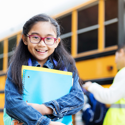 Filipino elementary age girl smiles as she waits to board school bus. She is holding books and a spiral notebook. The bus driver is in the background.