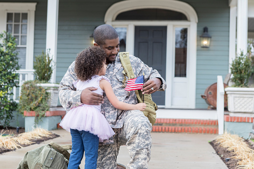 Preschool age girl hugs her soldier dad upon his return home from duty. They are holding a small American flag. They are on the sidewalk of their home.