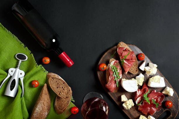 istock Red wine, cheese, cherry tomato, bread and prosciutto on wooden board over black backdrop, top view, copy space 950892886