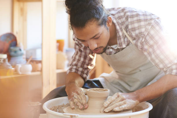 Craftsman making pottery ware Portrait of young handsome mixed race male potter making clay vase on spinning pottery wheel in workshop sculptor photos stock pictures, royalty-free photos & images