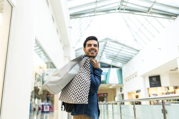 young man holding paper bags and walking in mall - shopping bag imagens e fotografias de stock