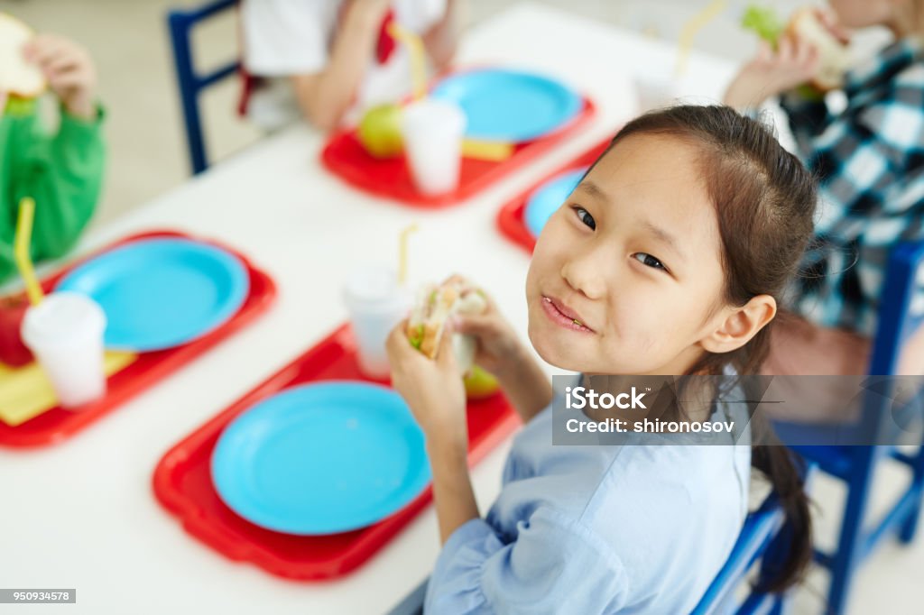 Happy girl eating school lunch Pretty Asian girl sitting at table with classmates in primary school canteen, eating sandwich and smiling at camera cheerfully Child Stock Photo