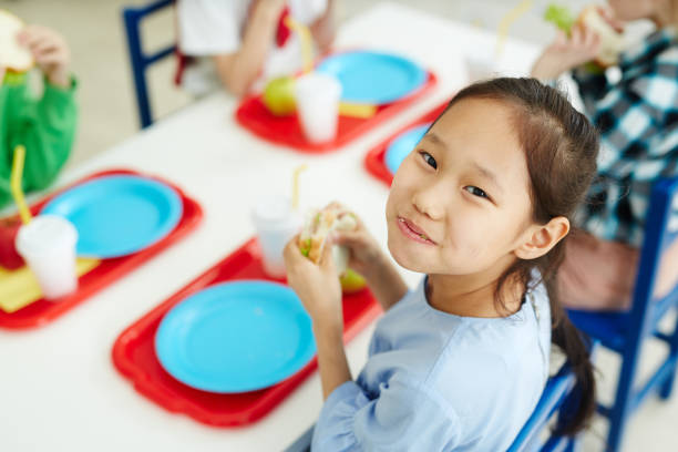 Happy girl eating school lunch Pretty Asian girl sitting at table with classmates in primary school canteen, eating sandwich and smiling at camera cheerfully school lunch child food lunch stock pictures, royalty-free photos & images