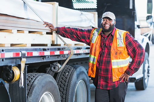 An African-American man in his 30s, standing next to a truck loaded with construction material. He is the truck driver delivering the supplies.