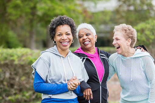 Three multi-ethnic women wearing hooded sweatshirts, hanging out together in the city, laughing while they take a walk. The woman with black hair, looking at the camera, is in her 50s and her senior friends are in their 60s.