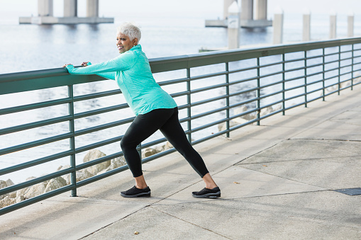A senior African-American woman in her 60s wearing sport clothing, exercising on a city waterfront. She is leaning on a railing, stretching her legs.