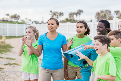 A group of multi-ethnic children and their teacher on a field trip by the sea. The teacher, a Pacific Islander woman in her 30s, and her students are standing next to a view finder, pointing and looking toward something in the distance.