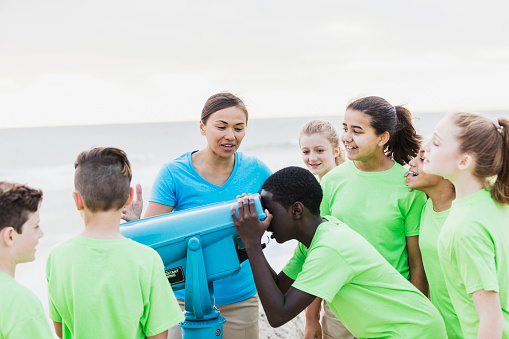A group of multi-ethnic children and their teacher on a field trip by the sea. The teacher, a Pacific Islander woman in her 30s, is talking to her students as an African-American boy looks through a view finder.