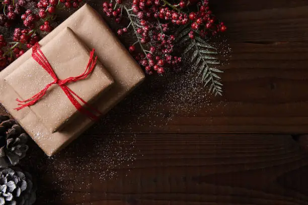 Top view of a plain brown paper Christmas present tied with red string, surrounded by pine cones and berries. Horizontal format with copy space.
