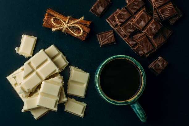 istock Cup of coffee, broken white and black chocolate bars and stack of cinnamon on black table, top view 950965968