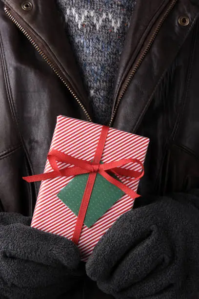 Closeup of a man wearing a sweater, gloves, and leather coat holding a wrapped Christmas present in front of his torso.