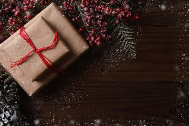 Top view of a plain brown paper Christmas present tied with red string, surrounded by pine cones and berries. Horizontal format with copy space and snow effect frame.