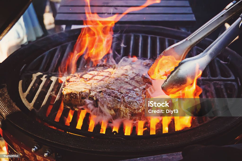 Steak on the grill with flames A piece of meat in flame. The cook flips a piece of meat beef on the grill with open fire. Steak on the grill with flames Barbecue - Meal Stock Photo