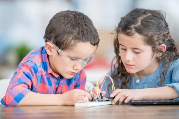 A young brother and sister are at school, working on a science project.  They are working with circuits and wires. The boy is wearing protective goggles.