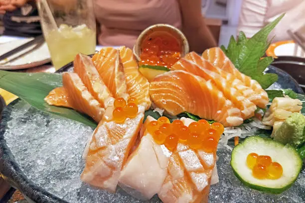 sashimi sliced salmon with salmon egg served on black ceramic stone bowl with full of ice for keep it fresh in restaurant environment with people on background