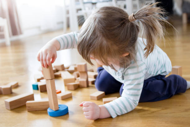 2 year old girl is playing and building a tower of wooden toy blocks 2 year old child is playing on the floor at home. She is building a tower of wooden toy blocks and are fully concentrated on balancing a piece on the top. playful set stock pictures, royalty-free photos & images