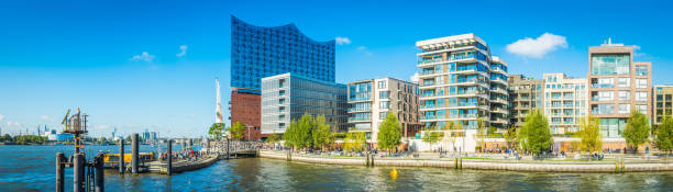 Panoramic view across the blue waters of the Norderelbe to the Elbphilharmonie Concert Hall, crowded promenade and modern apartment buildings of the redeveloped HafenCity quarter of Hamburg, Germany.