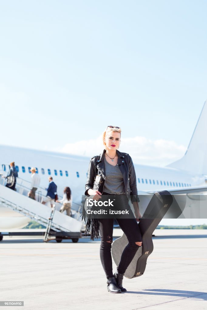 Female musician standing in front of an airplane Full length of young female musician standing in front of an airplane with a guitar. Woman rockstar going on vacation. Airport Stock Photo