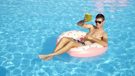 CLOSE UP Happy young guy on summer vacation lying on inflatable doughnut floatie in swimming pool & drinking pineapple alcohol beverage. Smiling man floating on inflatable pillow and sipping cocktail