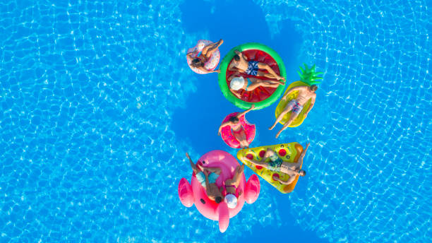 AERIAL: Cheerful teenage friends playing volleyball on fun floaties in pool AERIAL TOP DOWN COPY SPACE: Fit girls and muscular guys lying on colorful floaties in pool, playing volleyball with a ball. Friends on fun summer vacation floating on water on inflatable loungers pool party stock pictures, royalty-free photos & images