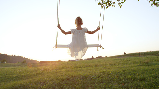 CLOSE UP: Young girl swinging on a wooden swing on a late summer evening. Little sister swaying outdoors gazing at the golden sunset. Playful daughter playing outside enjoying a beautiful sundown.