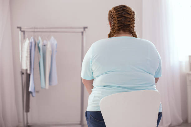 overweight young woman sitting on the chair New clothes. Overweight young woman sitting on the chair while thinking about new clothes excess fat stock pictures, royalty-free photos & images