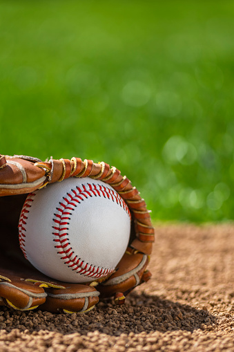 A low angle close-up view of a new baseball in a brown leather baseball glove sitting in the dirt with grass in the background