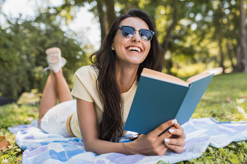 Beautiful portrait of a gorgeous young brunette woman reading a book in the park. Happy female student reading and learning a lesson on the book lying in the outdoor. Lifestyle, education, people.