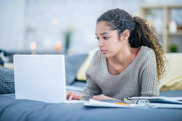 Doing Homework In Bed A teenage girl of African descent is indoors in her bedroom. She is laying in bed and typing on her laptop while doing homework. 13 14 years child teenage girls lying down stock pictures, royalty-free photos & images