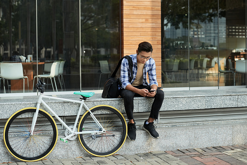 Full length portrait of handsome Asian cyclist sitting in front of modern coffeehouse and browsing Internet on smartphone