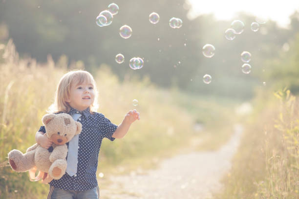 Little boy playing with soap bubbles and bear toy in park.