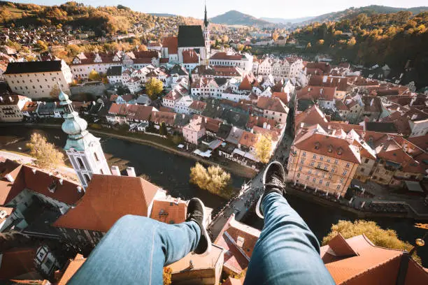 Aerial birds eye view of young rooftop climber sitting on the edge of a high rise tower dangling his feet high above the historic town of Cesky Krumlov, Bohemia, Czech Republic