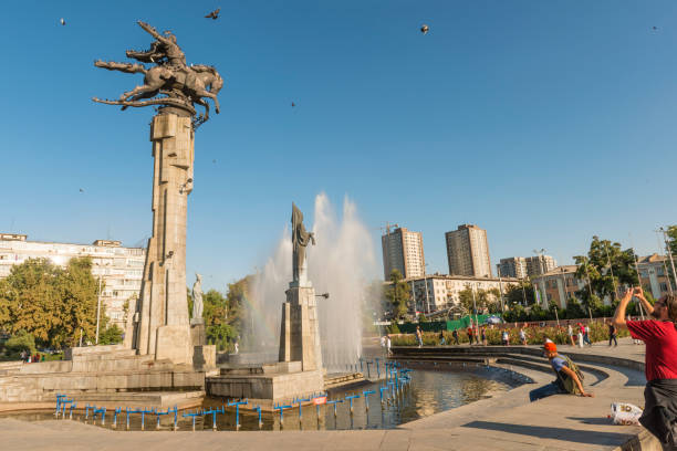 Manas Statue at the fountain, people taking photography and urban background on sunny day. Clear blue sky and some birds in the air. Bishkek, Kyrgyzstan