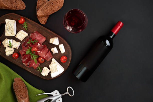 istock Red wine, cheese, cherry tomato, bread and prosciutto on wooden board over black backdrop, top view, copy space 951304470