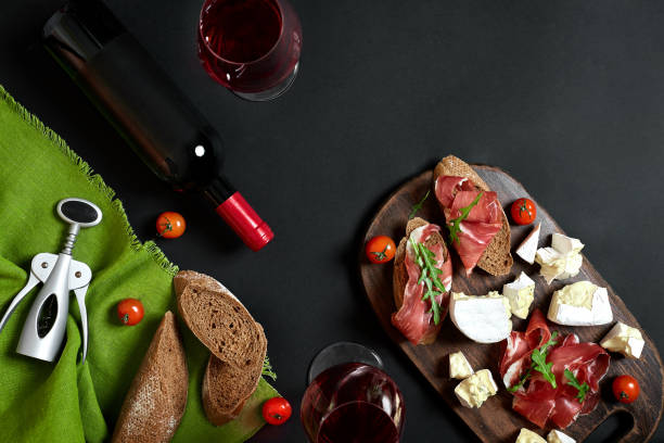 istock Red wine, cheese, cherry tomato, bread and prosciutto on wooden board over black backdrop, top view, copy space 951321584