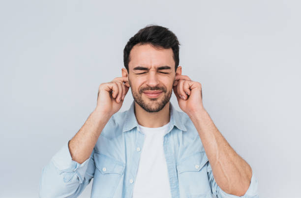 Horizontal studio portrait of handsome bearded young male plugs ears with displeased expression as hears annoying loud noise, isolated over white wall. People and emotion concept stock photo