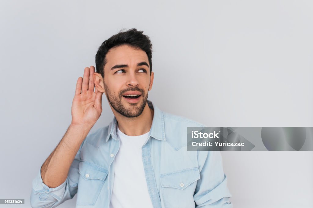 Portrait of handsome interested male placing hand on ear asking someone to speak up, isolated over white background. Young stylish man which overhears conversation in the studio. Copy space for text. Listening Stock Photo