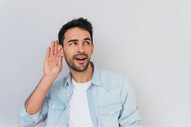 Portrait of handsome interested male placing hand on ear asking someone to speak up, isolated over white background. Young stylish man which overhears conversation in the studio. Copy space for text. stock photo
