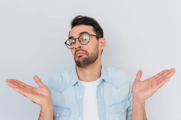 istock Caucasian unsure man make gestures doubtfully with hands with copy space. Uncertain young stylish stubble male with trendy round glasses wears demin blue shirt, shrugs shoulders being puzzled,confused 951331682