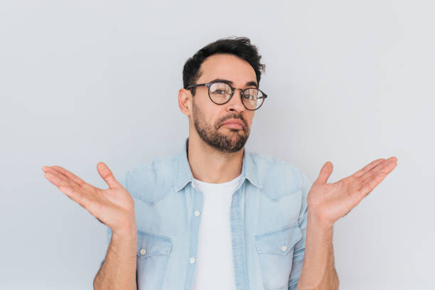 Portrait of uncertain young stylish stubble man with trendy round glasses wears demin blue shirt, shrugs shoulders being puzzled or confused. Caucasian unsure male make gestures doubtfully with hands. stock photo
