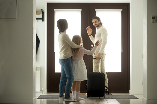 Smiling father waves goodbye to wife and daughter leaves home for business trip stands at door with travel suitcase, kid girl stays with mom seeing off dad moving out after divorce, family separation