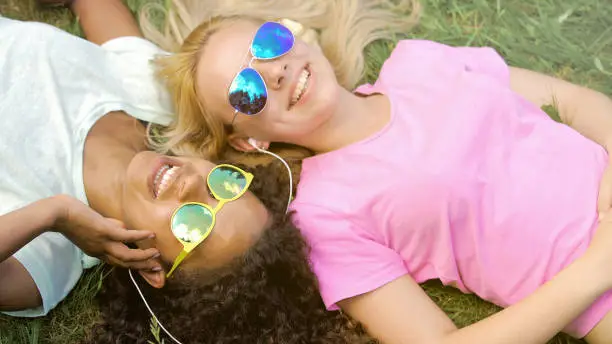 Two beautiful girls listening to music on smartphone, lying on grass on campus